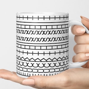 May include: A white ceramic mug featuring a repeating black graphic pattern of horizontal lines, geometric squares, interlocking loops, and triangular shapes arranged in distinct bands around the exterior surface of the cylindrical vessel.