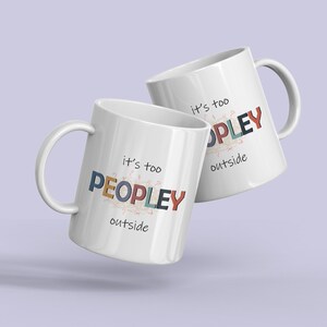 May include: Two white ceramic mugs with the text "it's too PEOPLEY outside" in colorful letters. The mugs have a curved handle and are set against a light purple background.