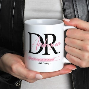 May include: White ceramic mug featuring the text "DR" in black, the name "Anderson" in pink script, and a pink loading bar graphic. Below the graphic is the text "LOADING...". The mug is held by a person wearing a black leather jacket.