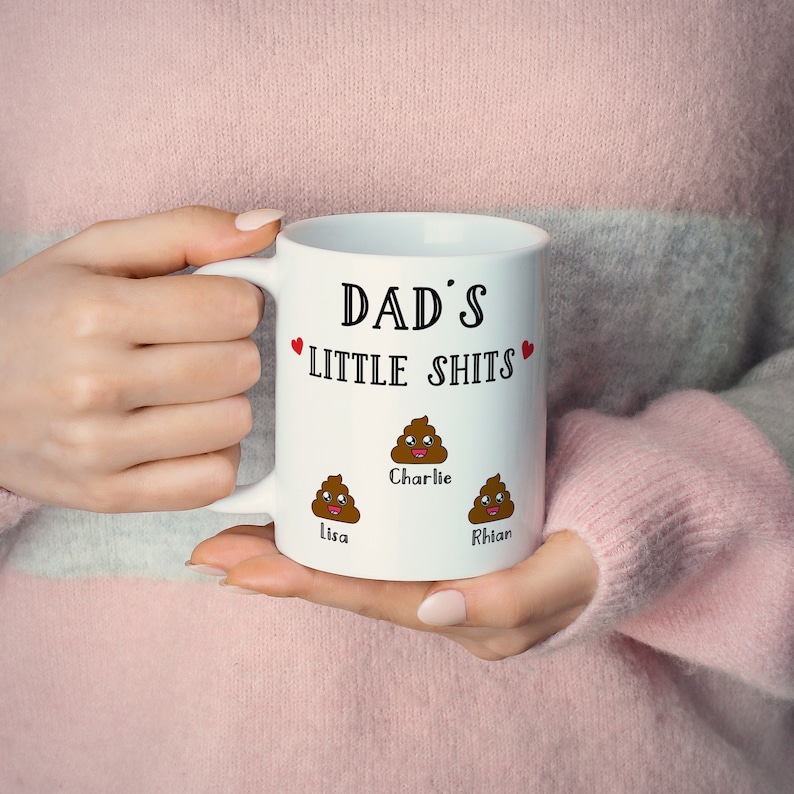 May include: A white ceramic mug featuring the black text DAD'S LITTLE SHITS with two small red hearts. Below the text are three brown cartoon poop emojis with smiling faces, each labeled with the names Charlie, Lisa, and Rhian in black.