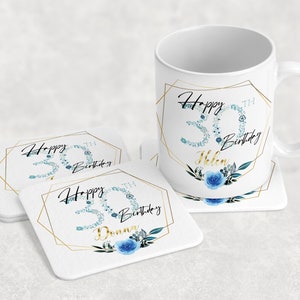 Może przedstawiać: Biały kubek ceramiczny z złotą geometryczną ramą i niebieskim wzorem kwiatowym. Kubek ma napis "Happy 30th Birthday Helen". Są dwa białe podkładki z tym samym wzorem, jeden z napisem "Happy 30th Birthday", a drugi z napisem "Happy 30th Birthday Donna".
