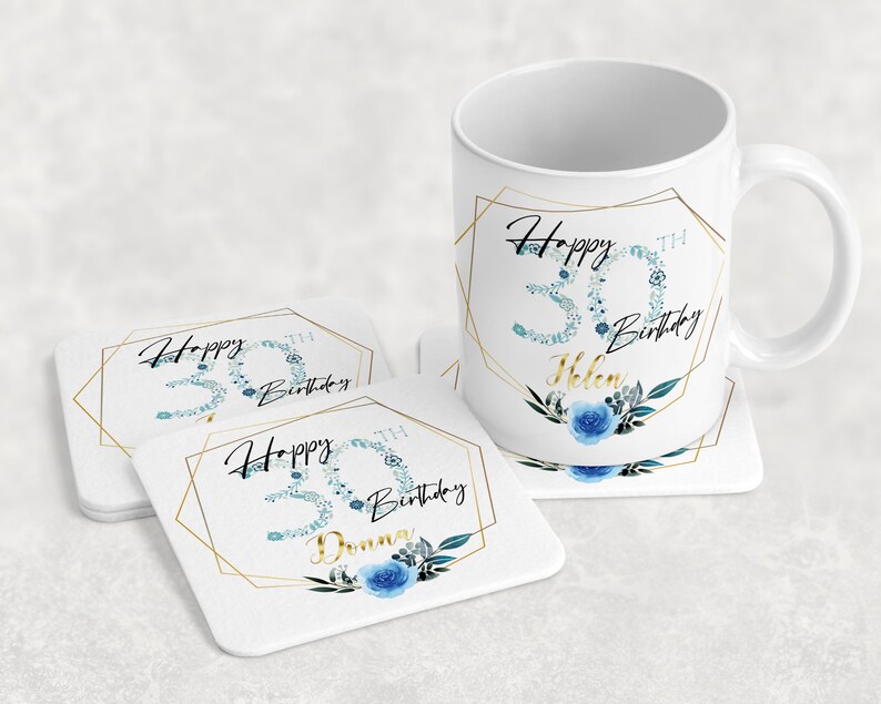 Może przedstawiać: Biały kubek ceramiczny z złotą geometryczną ramą i niebieskim wzorem kwiatowym. Kubek ma napis "Happy 30th Birthday Helen". Są dwa białe podkładki z tym samym wzorem, jeden z napisem "Happy 30th Birthday", a drugi z napisem "Happy 30th Birthday Donna".