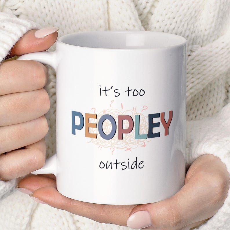 May include: White ceramic mug with the text "it's too PEOPLEY outside" in colorful letters. The mug has a handle and is held by a person wearing a white sweater. The mug is a standard 11 oz size.