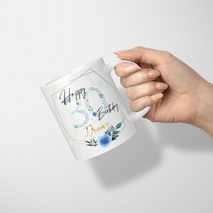 Może przedstawiać: Biały kubek ceramiczny z złotą geometryczną ramką i niebieskim wzorem kwiatowym. Kubek ma napis "Happy 30th Birthday Donna".