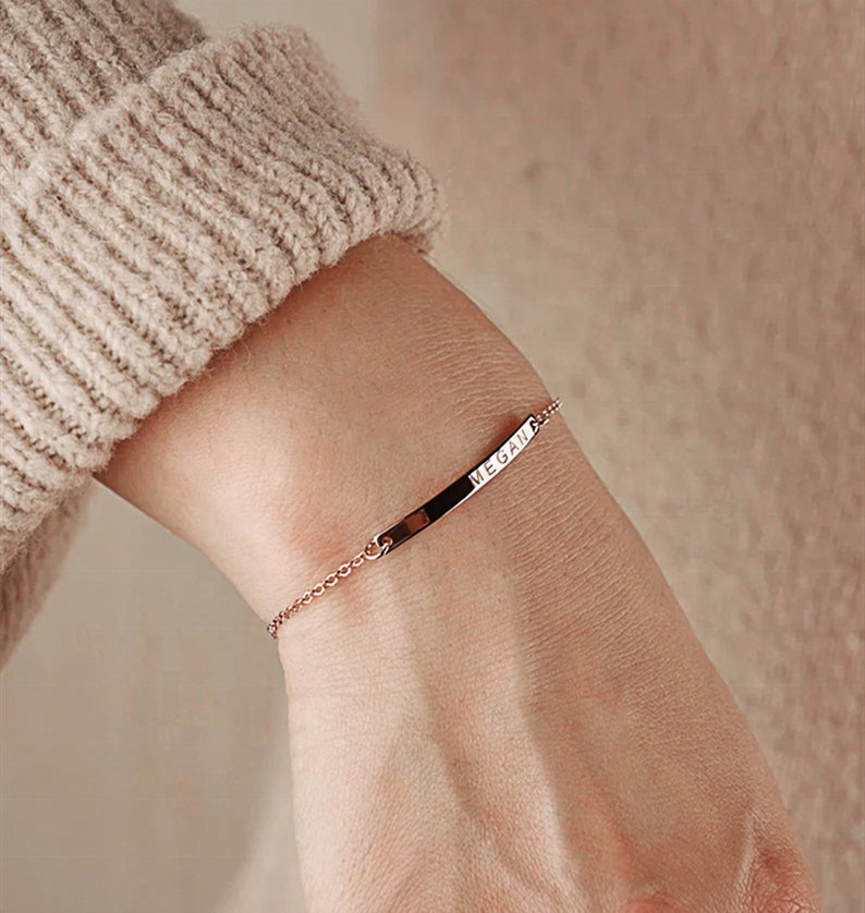 May include: A rose gold chain bracelet with a thin, rectangular bar charm engraved with the name "MEGAN".
