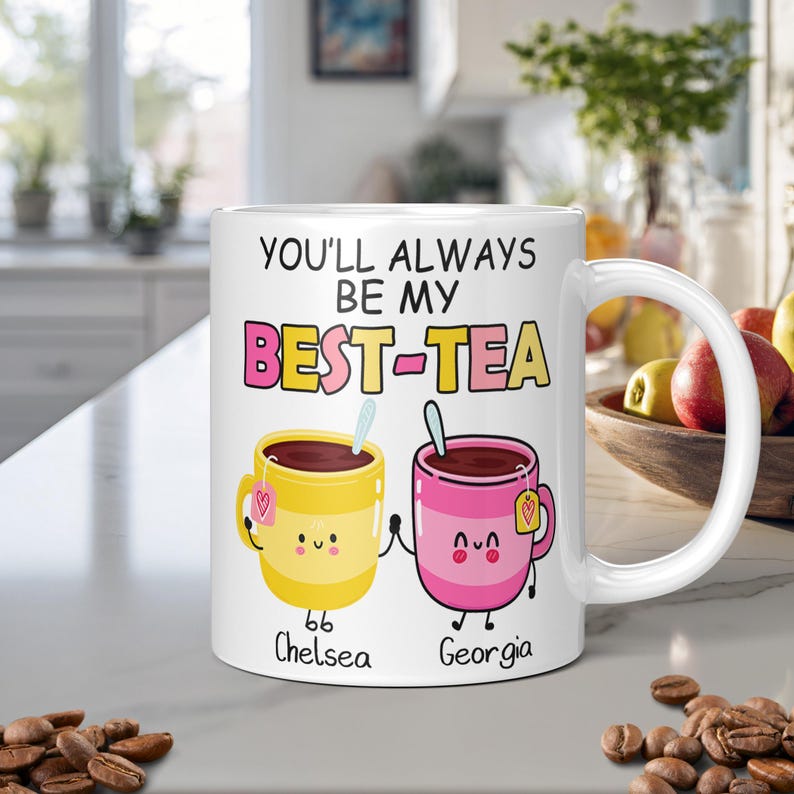 White personalised mug with the text "You'll always be my best-tea" and two cartoon teacups holding hands in a kitchen worktop with a kitchen on the background.