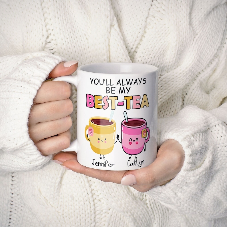 May include: White ceramic mug with the text "You'll always be my best-tea" and two cartoon teacups holding hands. The teacups are yellow and pink with smiling faces. The text "Jennifer" and "Caitlyn" are written below the teacups.