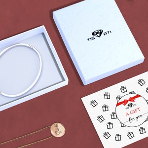 May include: A white gift box with a silver bangle inside. The box has the text "Tis Ati" on the top. A white card with the text "A Gift for you" and a red ribbon is on a red surface.