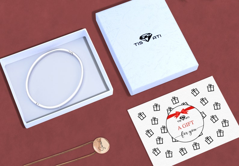 May include: A white gift box with a silver bangle inside. The box has the text "Tis Ati" on the top. A white card with the text "A Gift for you" and a red ribbon is on a red surface.