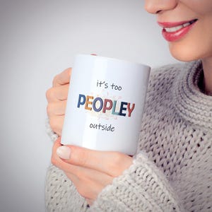 May include: White ceramic mug with the text "it's too PEOPLEY outside" in colorful letters. The mug is held by a person wearing a gray sweater. The mug is a standard size and shape, perfect for coffee or tea.