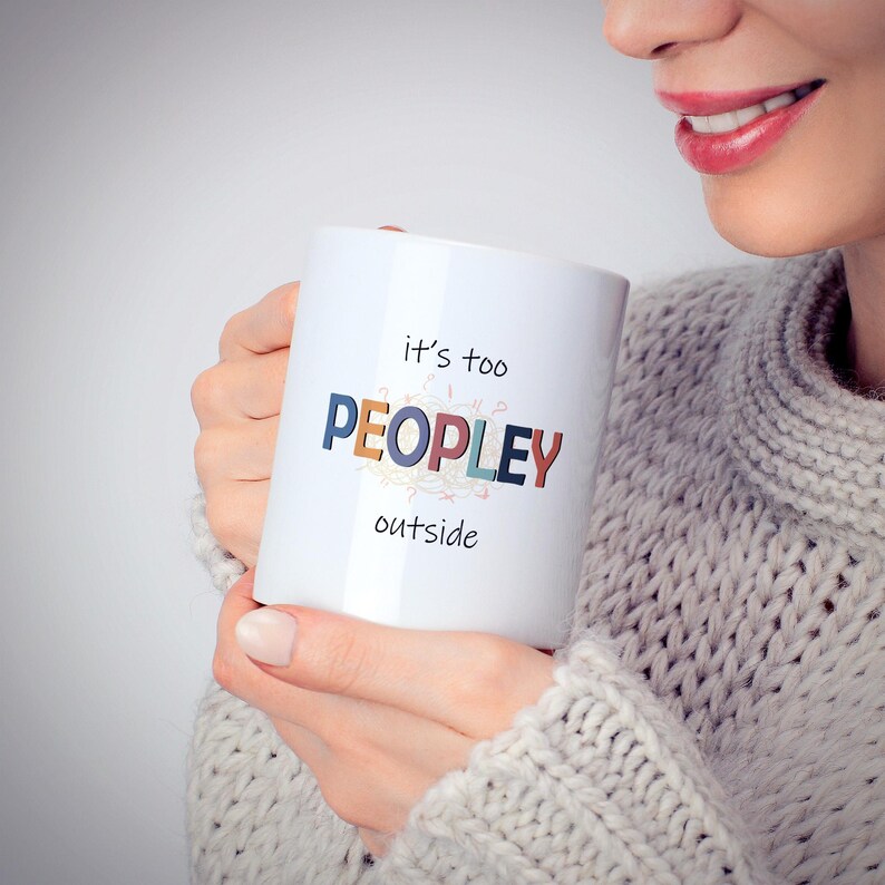 May include: White ceramic mug with the text "it's too PEOPLEY outside" in colorful letters. The mug is held by a person wearing a gray sweater. The mug is a standard size and shape, perfect for coffee or tea.