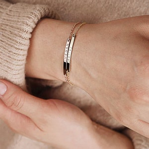 May include: A gold chain bracelet with two thin, rectangular charms. The charms are engraved with the names "Ashley" and "Megan".