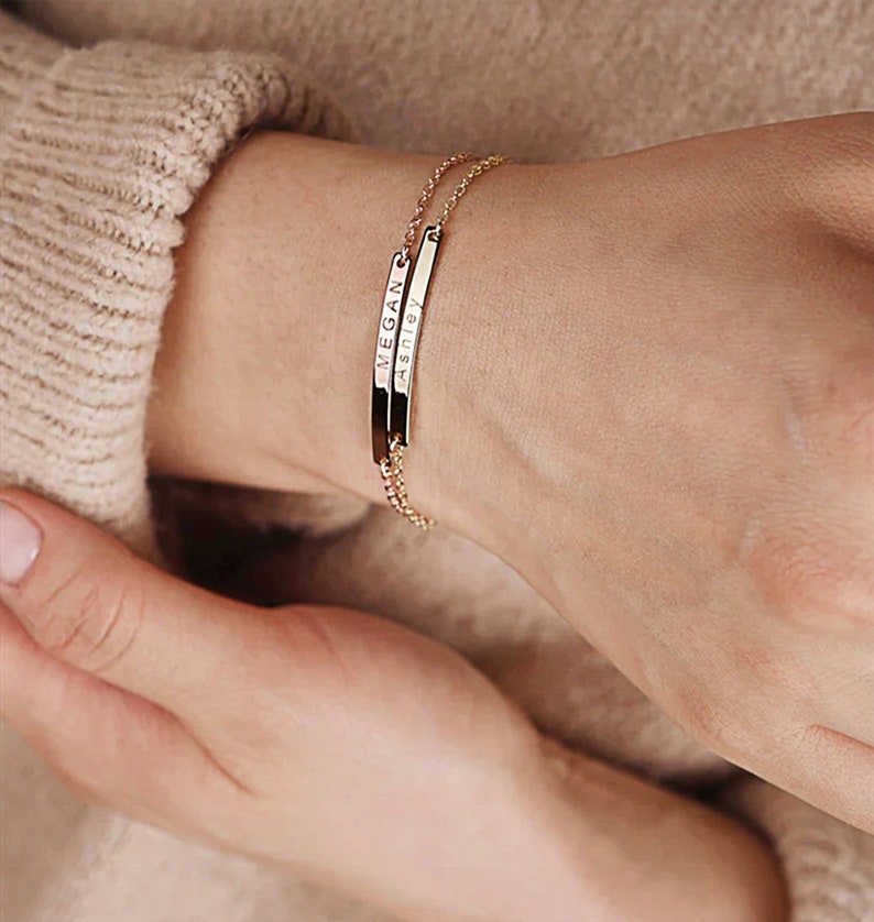 May include: A gold chain bracelet with two thin, rectangular charms. The charms are engraved with the names "Ashley" and "Megan".