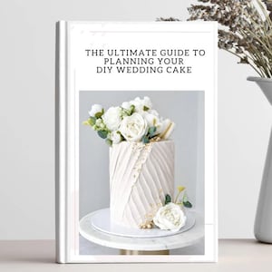 Op de afbeelding: Een wit boek getiteld "THE ULTIMATE GUIDE TO PLANNING YOUR DIY WEDDING CAKE" toont een versierde taart met witte bloemen. De taart staat op een witte taartstandaard. Een witte vaas met lavendel staat op de achtergrond.