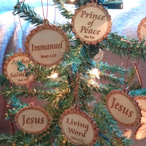 May include: Wooden Christmas tree ornaments with scalloped edges and the words "Immanuel," "Prince of Peace," "Jesus," and "Living Word" engraved on them. Each ornament has a scripture reference. The ornaments are hanging on a green Christmas tree.