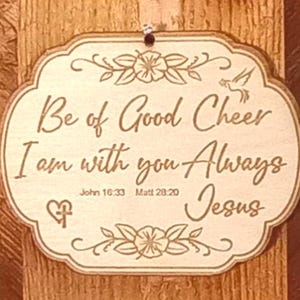 May include: A wooden plaque with the words "Be of Good Cheer, I am with you Always, Jesus" engraved in a decorative font. The plaque has floral and dove accents and includes the Bible verses John 16:33 and Matt 28:20.