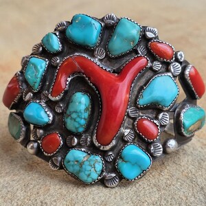 May include: A silver-toned cuff bracelet featuring a central red coral design surrounded by turquoise and red stones. The stones are set in a detailed, ornate setting, creating a striking contrast of colors and textures.