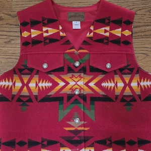 May include: A red wool vest with a Southwestern geometric pattern in black, yellow, white, and green. The vest has two front pockets, silver buttons, and a V-neck. The Pendleton label is visible.