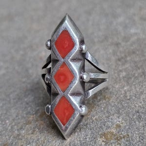 May include: A silver ring featuring three red diamond-shaped inlays. The ring has a long, pointed central design with small silver spheres and multiple bands. The ring is set against a neutral gray background.