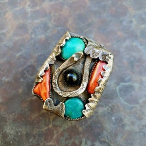 May include: A silver-toned ring with turquoise and coral accents. The ring features a coiled snake design, a black stone, and two turquoise stones. The ring has an ornate, textured design.