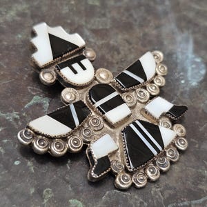 May include: A silver-toned, geometric brooch with inlaid black and white stones. The brooch features a central design with multiple sections, each with a unique shape and pattern. The stones are arranged in a symmetrical design, creating a striking contrast.