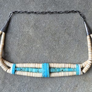 May include: A multi-strand necklace featuring cream-colored disc beads and a central section of bright turquoise disc beads. The necklace includes silver-toned metal cones at each end, connecting to a dark metal chain link closure.