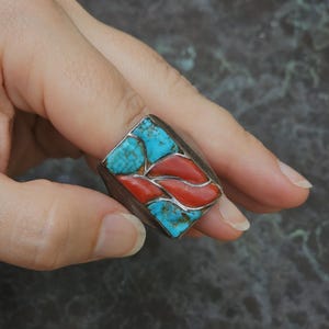 May include: A silver ring with a rectangular turquoise and coral inlay. The ring is set in a silver band and features a geometric design.