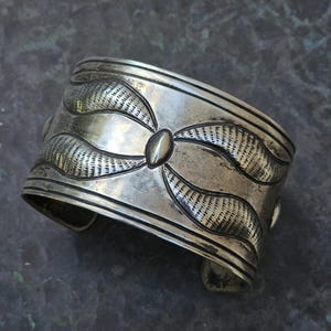 May include: A silver cuff bracelet with a geometric design. The bracelet is made of a thick, solid piece of silver and has a smooth, polished finish.