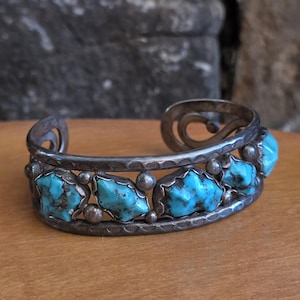 May include: Silver cuff bracelet featuring turquoise stones. The bracelet has a detailed design with a row of turquoise stones set in a silver frame. The turquoise stones are a vibrant blue color.