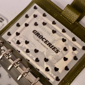 May include: An olive green planner with a clear overlay featuring black heart shapes. The overlay covers a US twenty-dollar bill with the word "GROCERIES" printed on it. The planner has a silver binder.