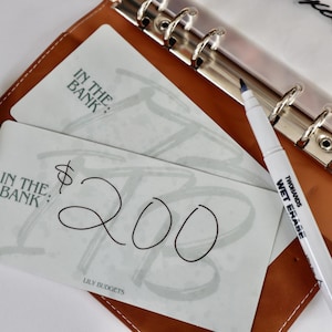 May include: Two white gift cards with the text "In the Bank" and a handwritten amount of $200. The cards are lying on a brown leather planner with a silver binder.