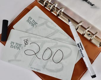 May include: Two white gift cards with the text "In the Bank" and a handwritten amount of $200. The cards are lying on a brown leather planner with a silver binder.