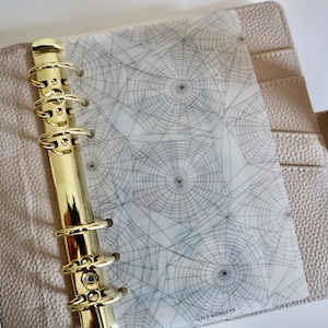 May include: A beige textured leather binder featuring a gold-toned six-ring metal mechanism and a translucent vellum dashboard insert printed with a black spiderweb pattern and the text LILY BUDGETS at the bottom right corner.