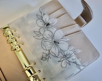 May include: A beige textured personal planner with a gold six-ring binder mechanism. A translucent vellum dashboard insert features a black line art floral illustration and the text LILY BUDGETS printed in the bottom right corner.