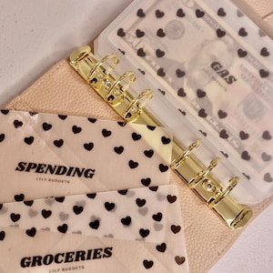 May include: A light pink budget binder with gold-colored rings and clear plastic dividers. The dividers are decorated with black heart patterns. The words "SPENDING" and "GROCERIES" are printed on the dividers.
