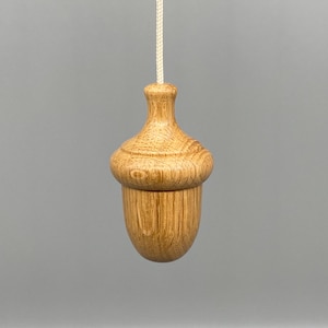 May include: A wooden acorn light pull with a natural wood grain. The pull is suspended by a cream cord against a grey background. The acorn design adds a touch of nature-inspired decor, suitable for any room.