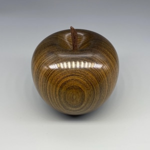 May include: A polished wooden apple sculpture. The apple is a warm brown colour with darker wood grain patterns. A small, brown stem protrudes from the top. The sculpture is a decorative item, suitable for display.