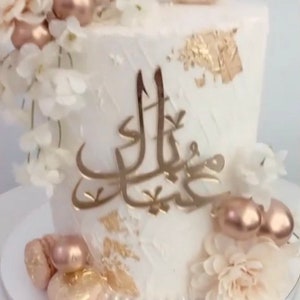May include: A white cake decorated with gold leaf, white flowers, and a gold Arabic inscription. The cake is topped with a white crescent moon and a cluster of rose gold balloons.