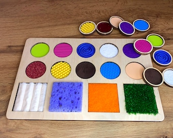 Puede incluir: Un tablero sensorial de madera con varias texturas y colores. El tablero tiene 12 recortes redondos con diferentes texturas, incluyendo tela, espuma y madera. También hay cuatro recortes cuadrados con diferentes texturas, incluyendo tela, espuma y hierba. El tablero está diseñado para ayudar a los niños a desarrollar sus sentidos táctiles.