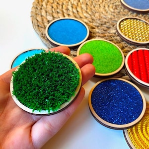 Puede incluir: Una colección de discos sensoriales con diversas texturas y colores. Un disco presenta césped artificial verde, mientras que otros muestran texturas azules, verdes, rojas y amarillas. Cada disco está enmarcado en madera.
