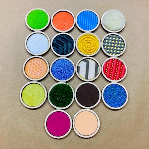 May include: A set of 16 round wooden discs with different textures and patterns. The discs are arranged in a grid pattern. The textures include a variety of fabrics, materials, and patterns, such as stripes, polka dots, and swirls.