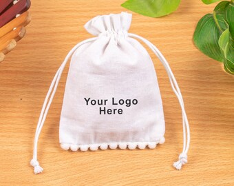 May include: A white drawstring bag with the text "Your Logo Here" printed on the front. The bag has a pom-pom trim along the bottom edge and white drawstrings. The bag is made of a textured fabric.