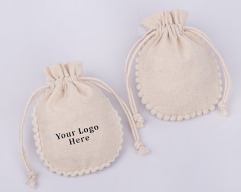 May include: Two small, off-white cotton drawstring bags with white pom-pom trim. One bag has the text "Your Logo Here" printed on the front.