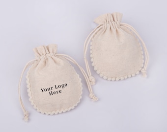 May include: Two white cotton drawstring bags with a white pom-pom trim. One bag has the text "Your Logo Here" printed on the front.