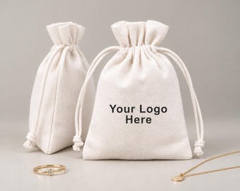 May include: Two off-white drawstring pouches made of fabric, with the text Your Logo Here printed in black on the front of the larger bag. A thin gold ring with a small clear stone and a delicate gold chain necklace are placed in front.