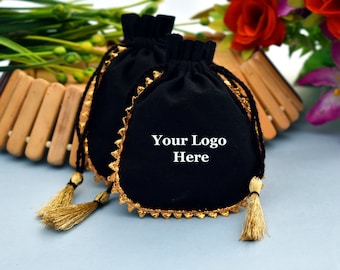 Könnte beinhalten: Zwei schwarze Samt-Kordelzugbeutel mit goldfarbenem Besatz und Quasten. Auf den Beuteln steht der Text "Your Logo Here". Die Beutel liegen auf einer hellblauen Oberfläche mit Blumen im Hintergrund.