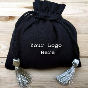 May include: A black drawstring pouch with silver tassels. The pouch has the text "Your Logo Here" in white. The bag is made of a soft fabric and is tied with a black rope.