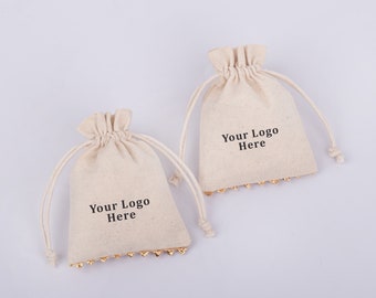May include: Two small, white cotton drawstring bags with gold trim. Both bags have the text "Your Logo Here" printed on the front.