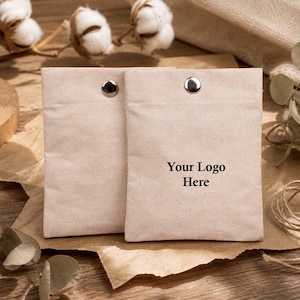 Puede incluir: Dos bolsas de lona beige con cierres de botón a presión plateados. El texto "Your Logo Here" está impreso en la parte delantera de una bolsa. Las bolsas están sobre una superficie de papel marrón, con capullos de algodón y cuerda en el fondo.