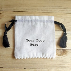 May include: White cotton drawstring bag with black tassels and a white pom-pom trim. The bag has the text "Your Logo Here" printed on the front.